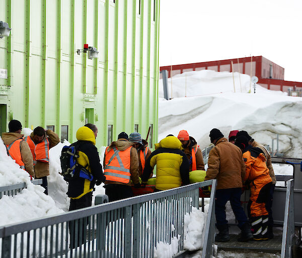 Line of expeditioners pass a stretcher from the building into the back of a Hagglunds vehicle.