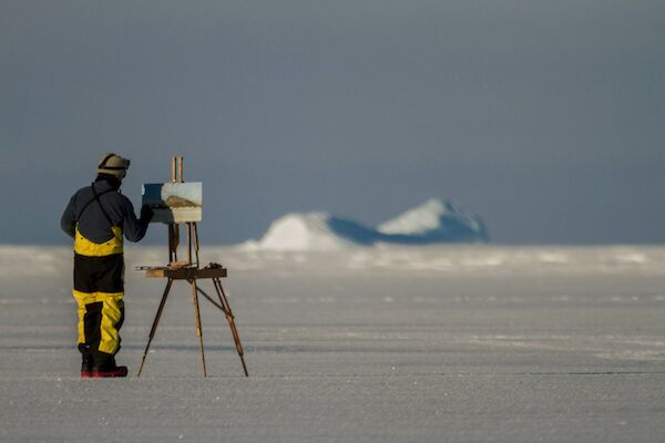 JOhn Kelly wearing protective Antarctica gear, paints with an easel set up on the ice,
