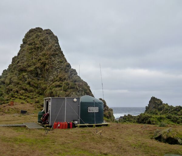 A water tank hut with an annex, aerial, decking and fuel containers on a grassy area with rock stacks and the west coast in the background
