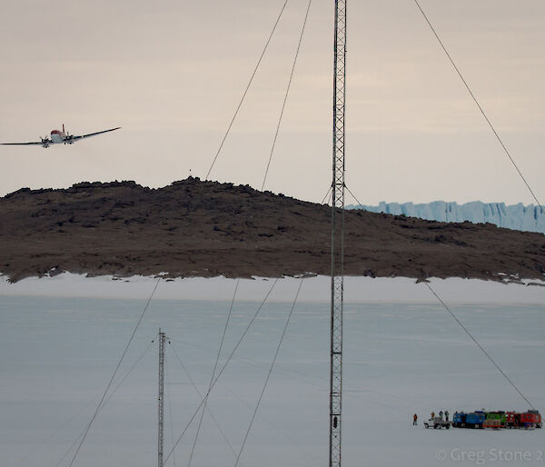 The Basler plane is seen to approach the Mawson Ski Landing Area from beyond Bechervaise Island. The ground crew consisting of the Mawson Emergency Response Team and Refueling Team along with the two passengers bound for Davis can be seen gathered on the sea ice in the foreground