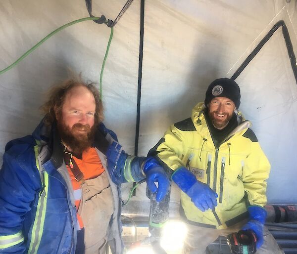 Two men stand in a tent with a drill.