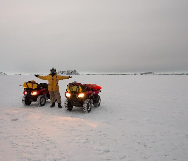 Daniel standing with two Quad bikes before sunrise on the sea ice with shore in the distance