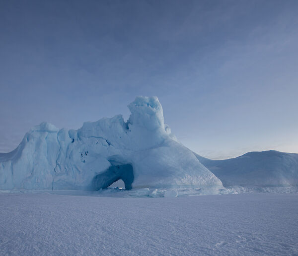 An ice burg locked in the sea ice that has a large hole weathered in the side.
