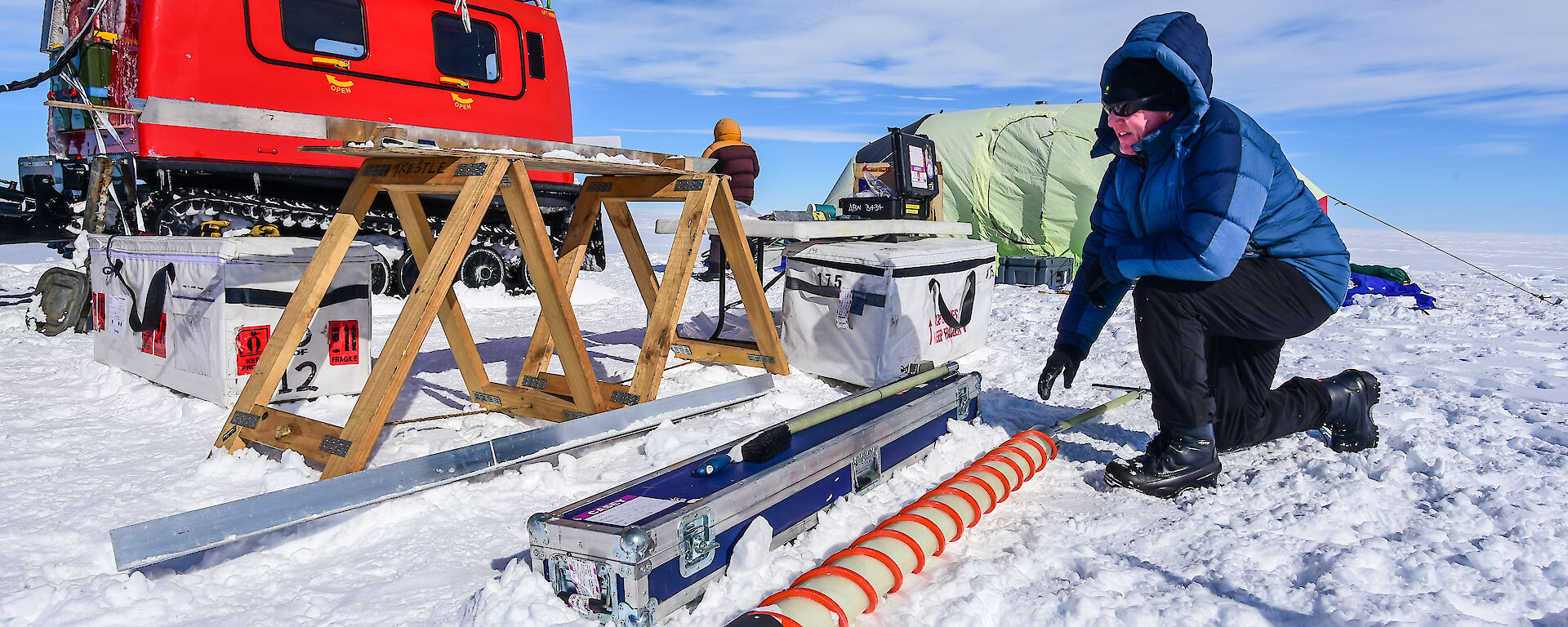 A scientist extracts ice from the ice core drill, with the campsite and Hägglunds in the background.