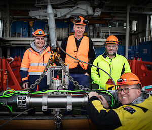 4 crew members stand with a piece of scientific equipment that is attached to large chains ready to be deployed from a ship.