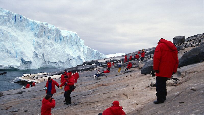 Tourists in various poses are dotted on shore with a glacier in background. Most of them are wearing the same coat.