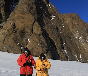 Two expeditioners stand in front of a small peak and use handheld GPS