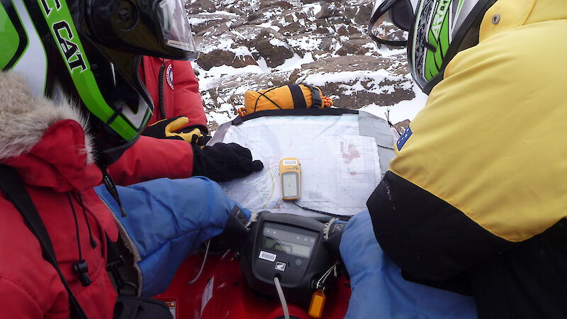 A GPS unit and map lie in the middle of a quad bike, while two expeditioners use them for navigation at a stop.