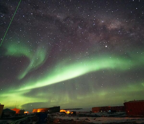 A green laser probes high-altitude clouds above Davis research station, in a night sky of auroras and stars.