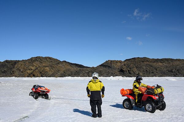 A man stands beside another man on a quad bike as it is being pulled out of the ice