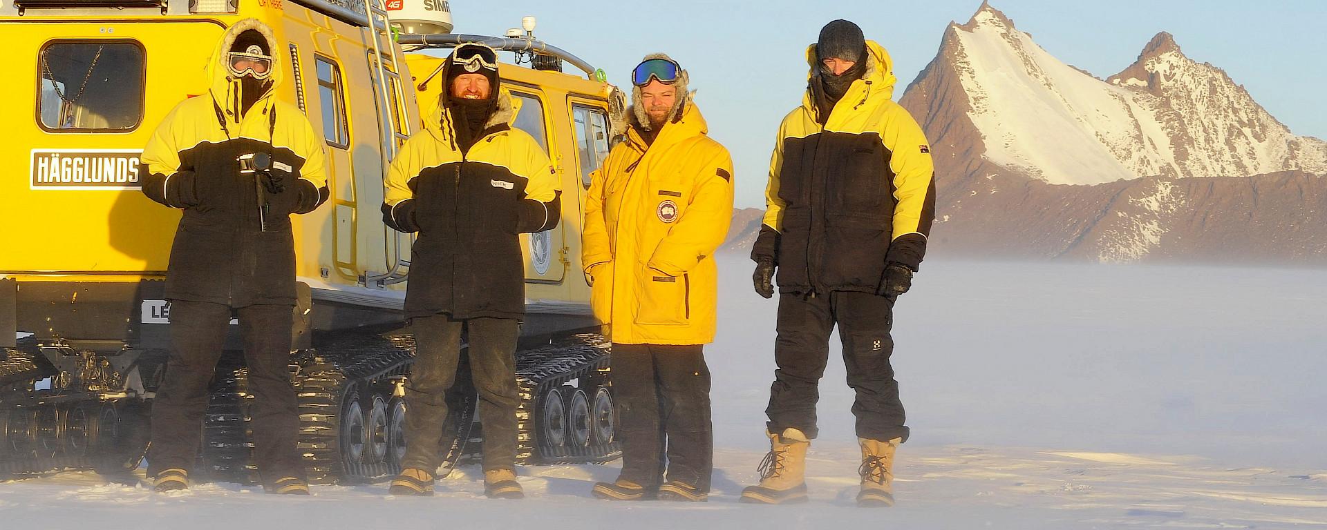 Expeditioners stand in front of a yellow Hägglunds in a snowy landscape.