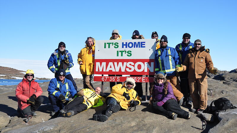 A group of expeditioners surround a sign at Mawson that reads “It’s home, it’s Mawson”