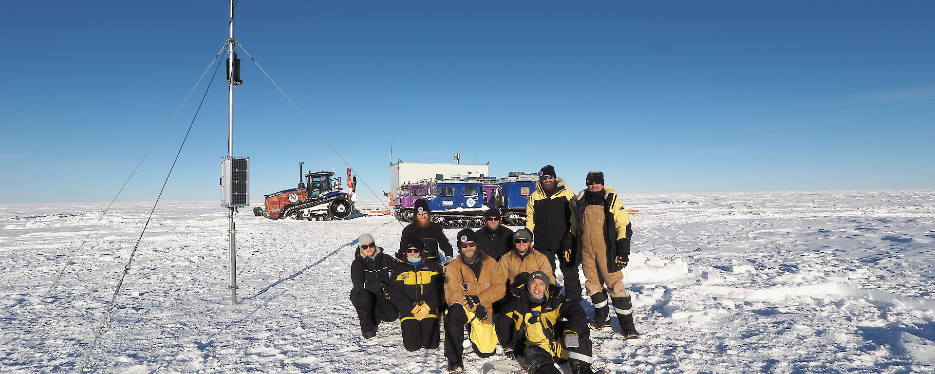 A group of expeditioners pose in an icy landscape with colourful vehicles in the distance.
