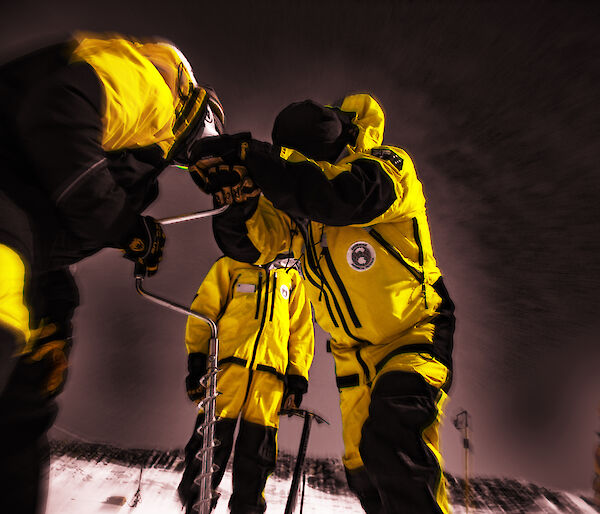 Three people wearing yellow jackets holding a piece of equipment with a dark sky behind them.