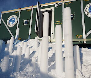 Cylinders of ice stand vertically in the snow outside a green container.