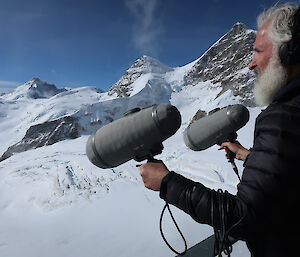 A man holding two long microphones pointed towards a snowy mountain range