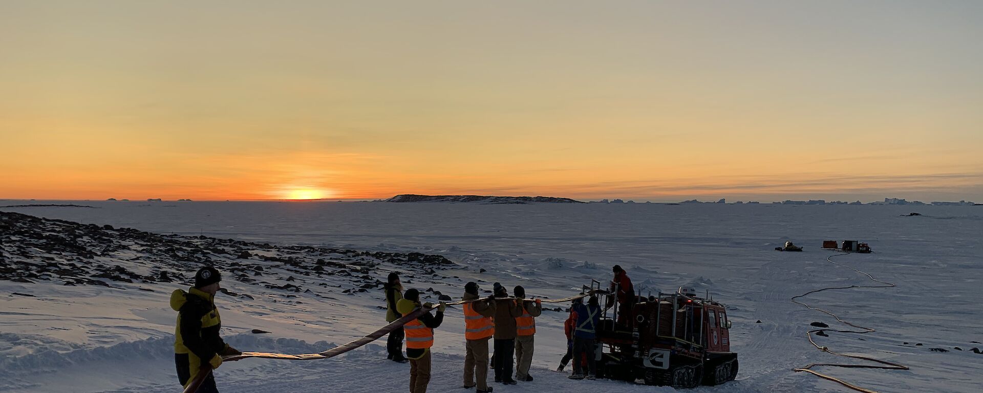 People in a line holding a hose being fed into a hagglund on the ice, with a sunset on the horizon.