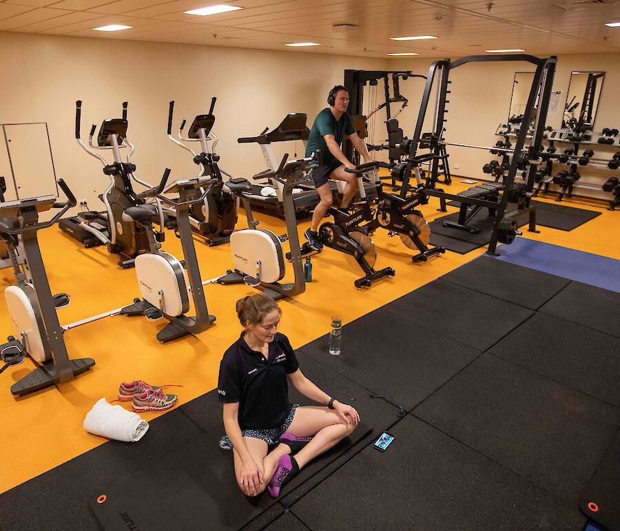A woman sitting on a yoga mat and a man pedalling an exercise bike, surrounded by other bikes and gym equipment.