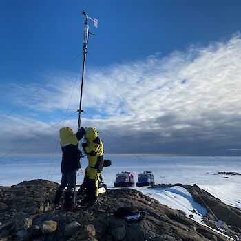 Expeditioners fixing an automated weather station with Hägglunds oversnow vehicles in the background