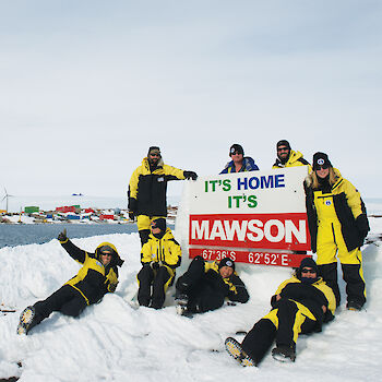 Expeditioners posing with the Mawson sign on West Arm. Station in background.
