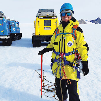 Expeditioner in climbing gear in front of two oversnow vehicles.