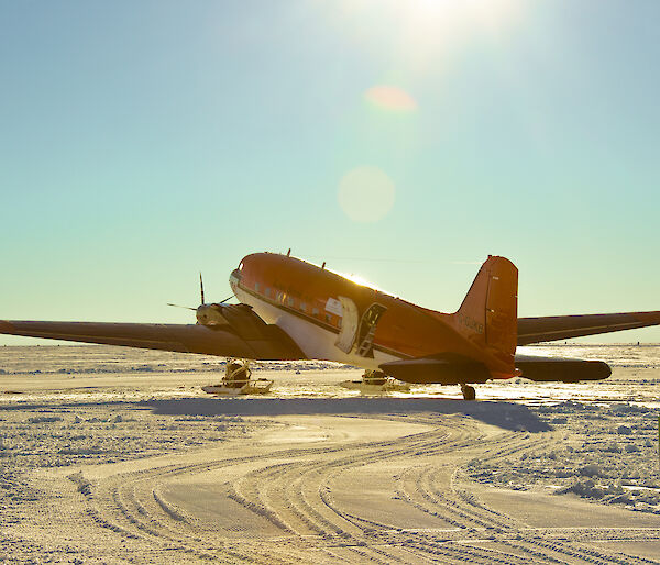 A Basler aircraft, bright orange with a white underside and ski apparatus fixed to its front wheels, is parked on a flat, snowy plain under a clear blue sky