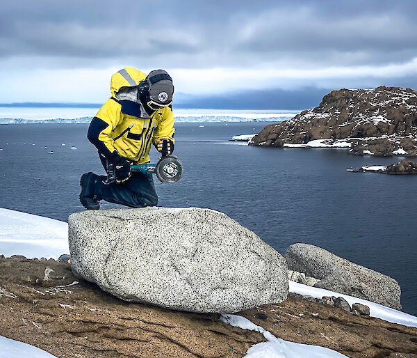 A woman leans against a rock with an angle grinder with the ocean and ice behind her