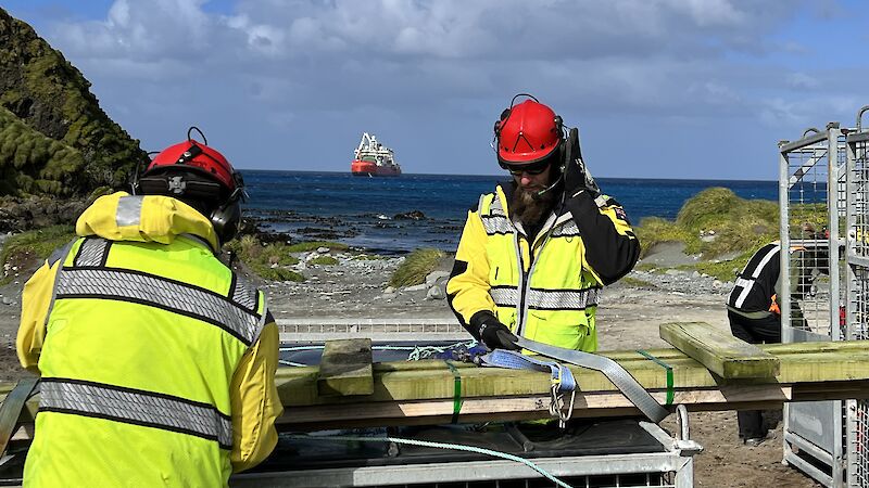 2 expeditioners in hi-vis examine cargo in a metal crate. An orange and white ship floats in blue water in the distance.