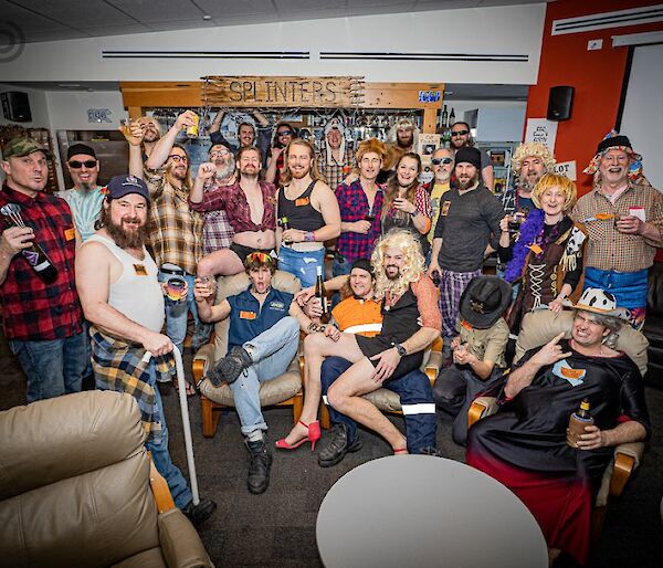 A group of about 25 people, many of them in checked flannel shirts, some wearing blonde, curly wigs, assembled close together in a lounge area for a group photo