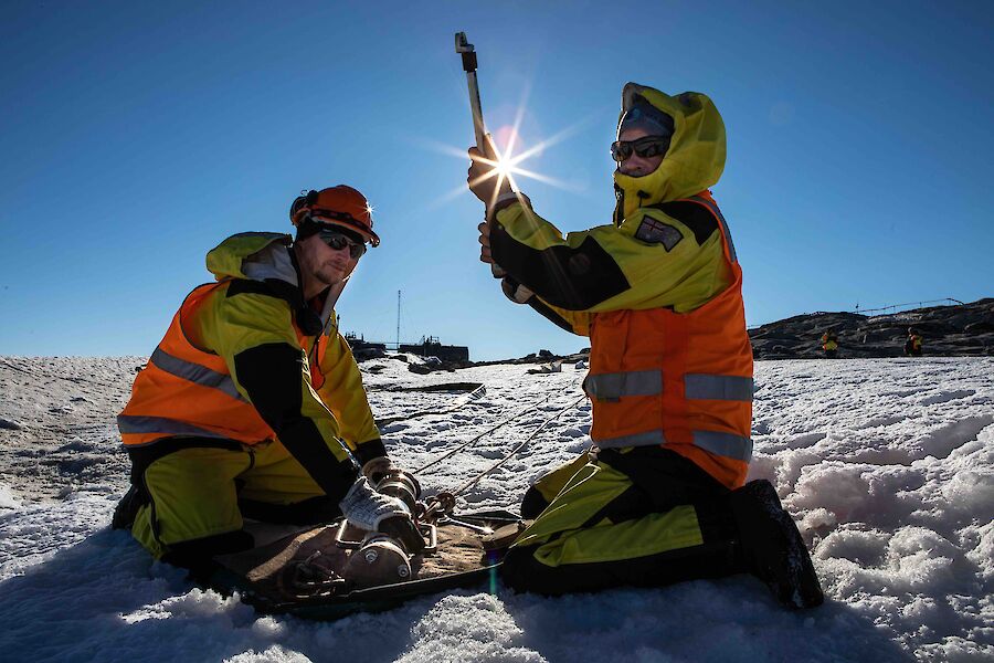Two men in hi vis sitting on the ice holding refueling equipment