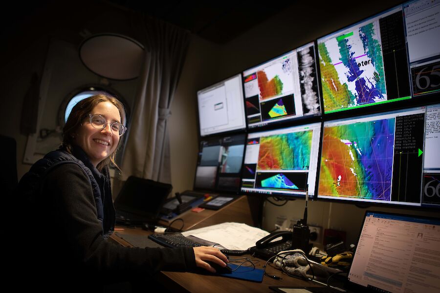 A woman sits at a desk in front of a wall of screens depicting colourful echosounder images.  She smiles to camera.
