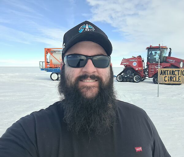 A man in black with a beard stands on ice in front of the Antarctic Circle sign, with a tractor in the background