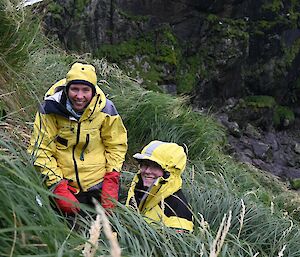 Two people in yellow outdoor gear smiling on a tussocky hillside