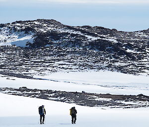 Two people walk across a snowy slope with a rocky hill in the background.