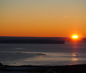 The sun sinks below the horizon, with an icy sea in the foreground.