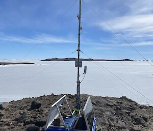 A portable weather station secured to a rock outcrop in the foreground. Sea ice and blue sky with landscape in the background