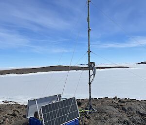 A portable weather station secured to a rock outcrop in the foreground. Sea ice and blue sky with landscape in background