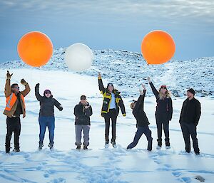 Seven expeditioners from the Bureau of Meteorology pose for a photo holding some weather balloons within Casey station a snowy hill pictured in background