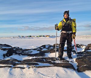 A man in yellow outdoor gear stands, with two walking poles, on rocks surrounded by snow.