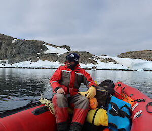 A man in red survival gear sits in a red rubber dingy on a still bay with snow and rocks behind him