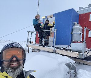 A selfie of Adam and expeditioners outside a red hut blowing snow with smiles