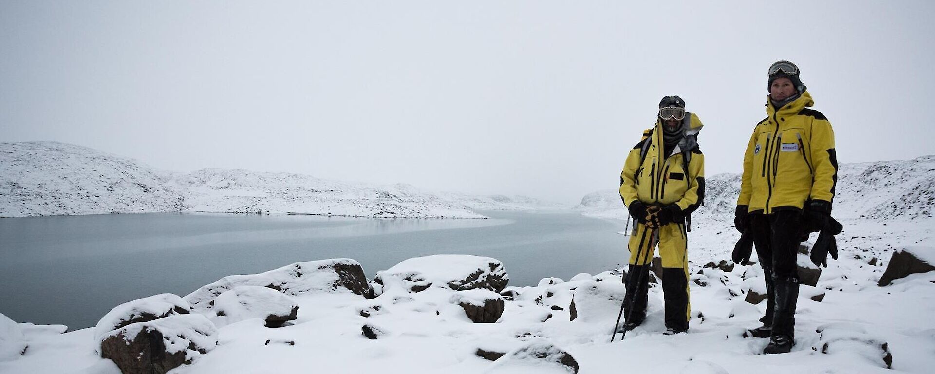 Two expeditioners dressed in yellow wind proof clothing with the snow covered hills and Deep Lake in the background.