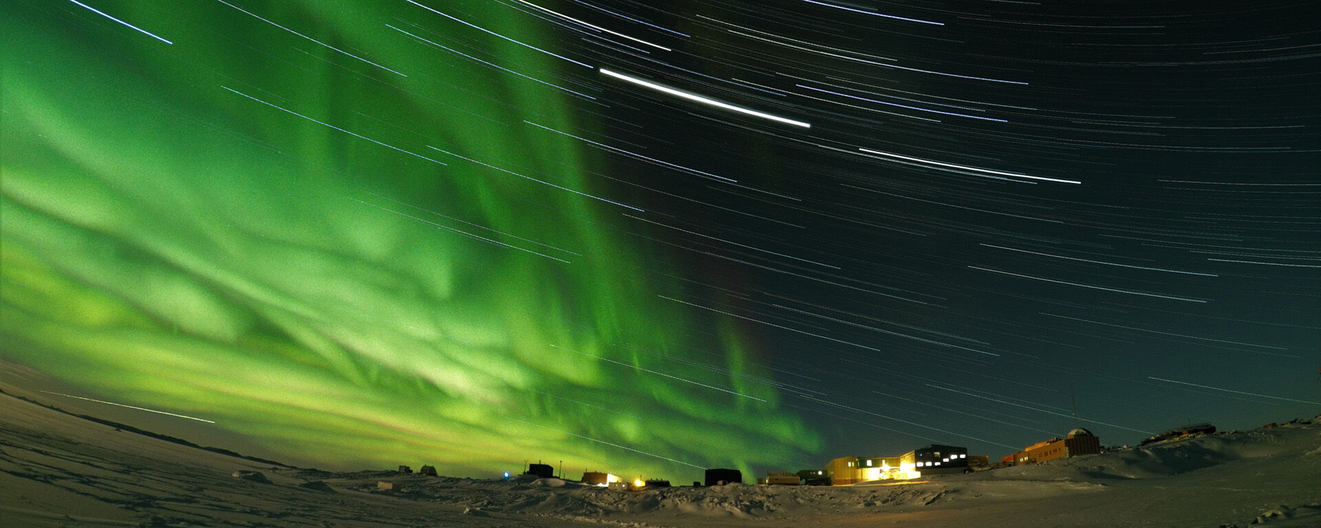 Long exposure at night with star trails and green Aurora lights, in the distance buildings have lights on.