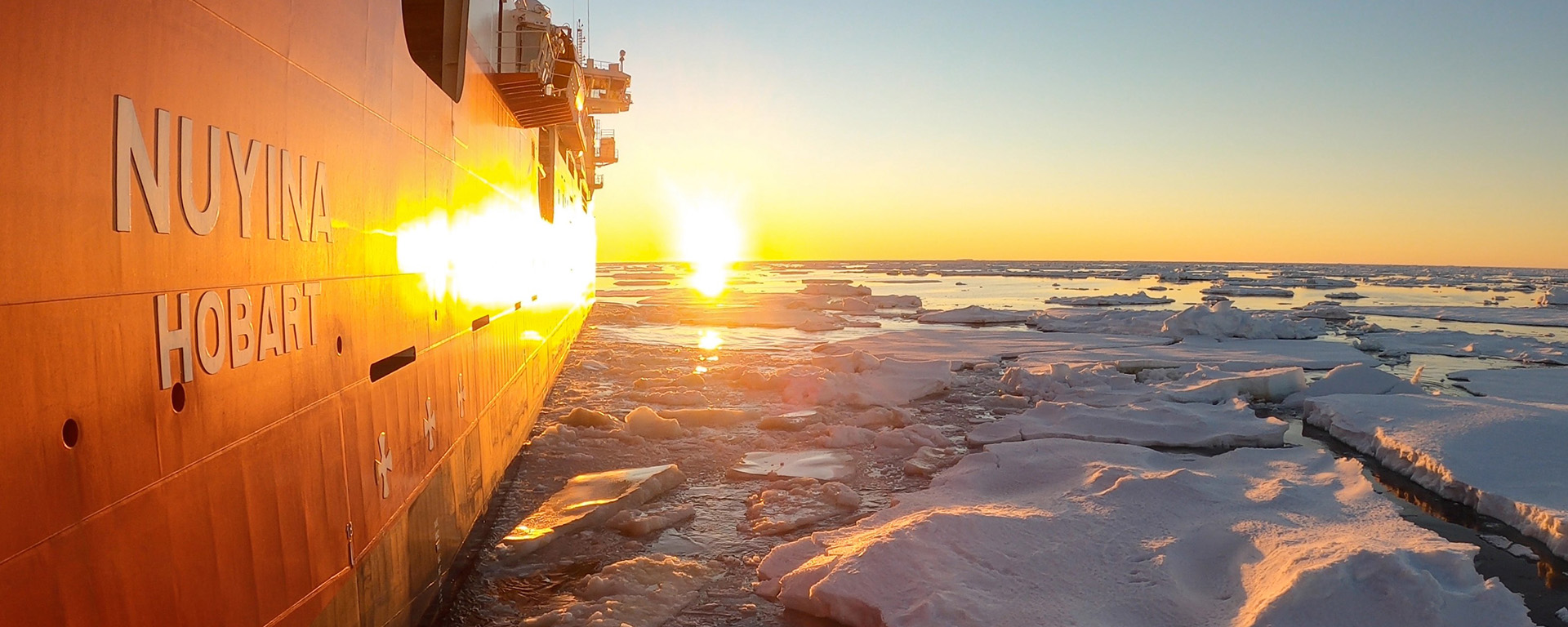 An orange and white ship sails through the ocean, surrounded by floating chunks of ice. The sun is setting in a clear sky.