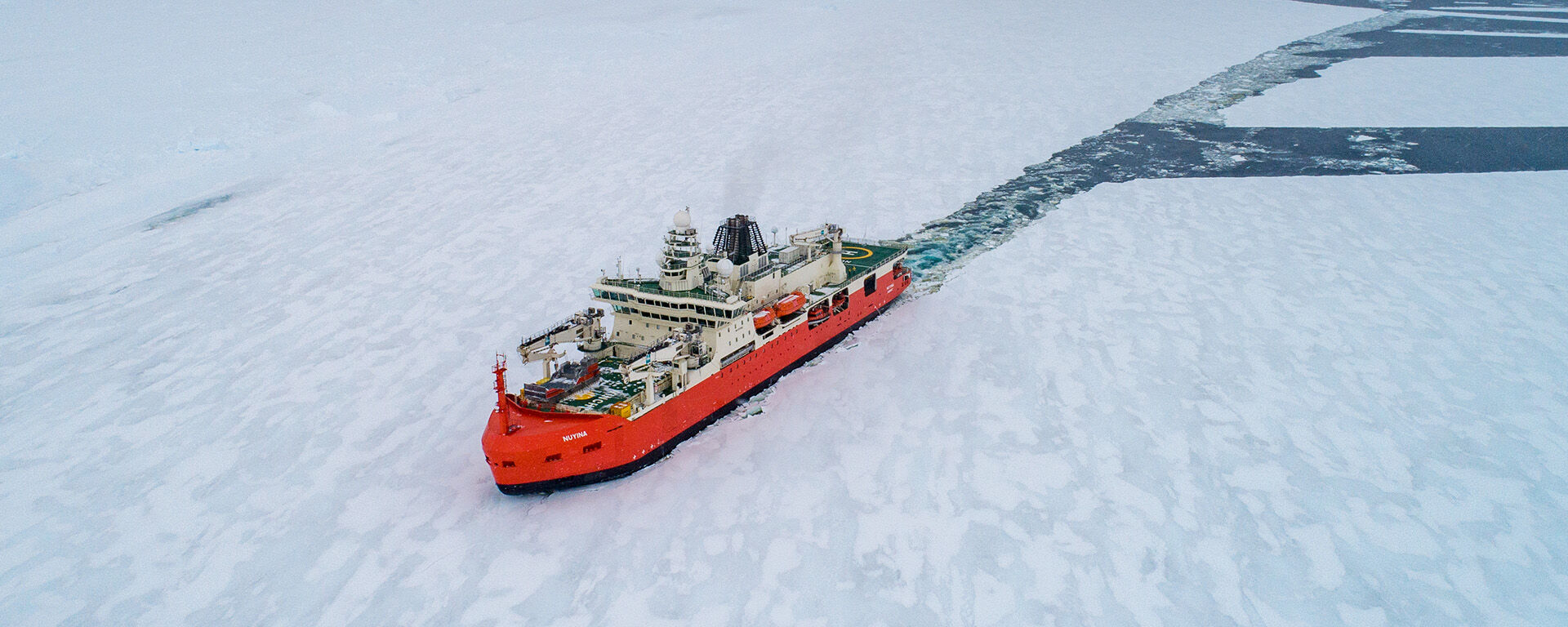 An aerial drone photo of an orange and white ship breaking through sea ice. There is a long channel of water behind the ship where it has broken a path through.