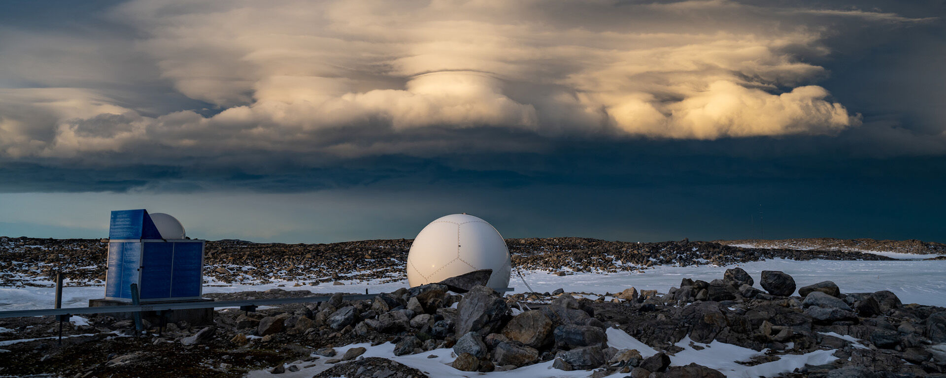 Large, illuminated clouds roll in over an icy and rocky landscape. There are a couple of buildings in the foreground.