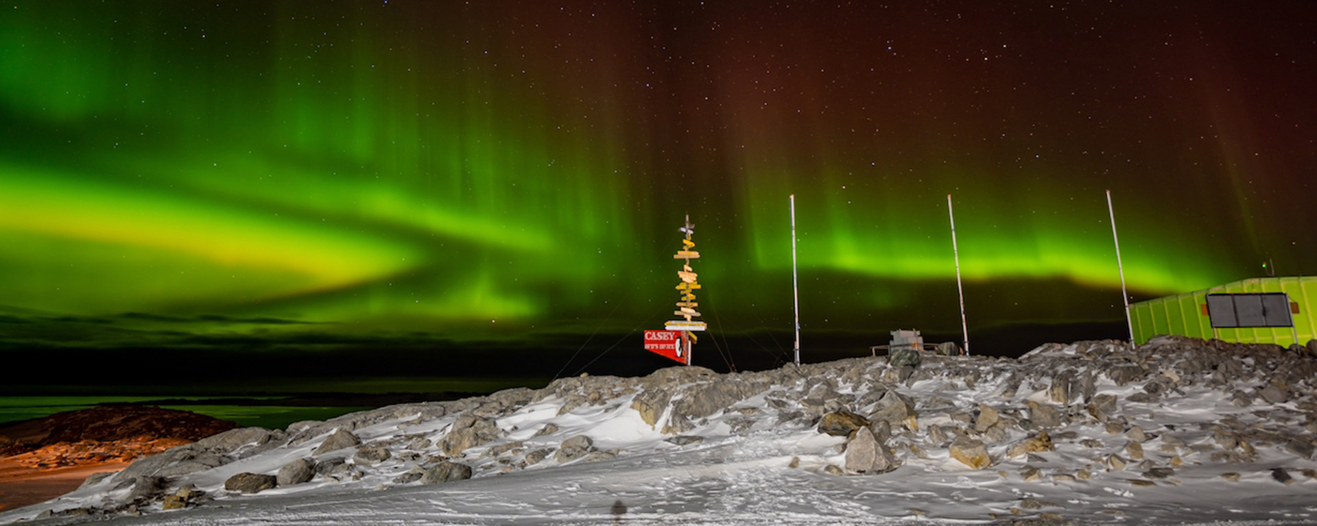The Aurora Australis in the sky above a signpost, flagpoles and a shed. It appears like lakes and bands of green light, fading through dull red on their upper edges into a starry night sky.