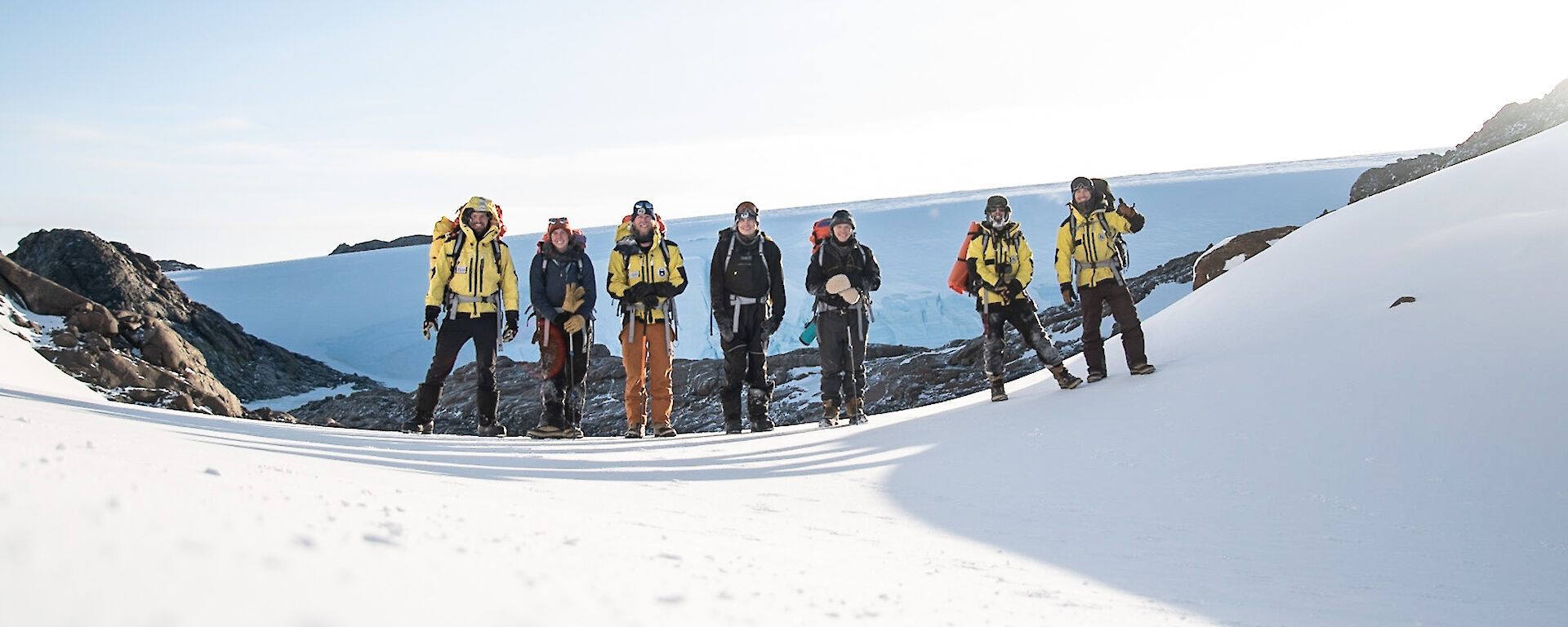 Seven people wearing snow gear standing in a row in a snow covered landscape.