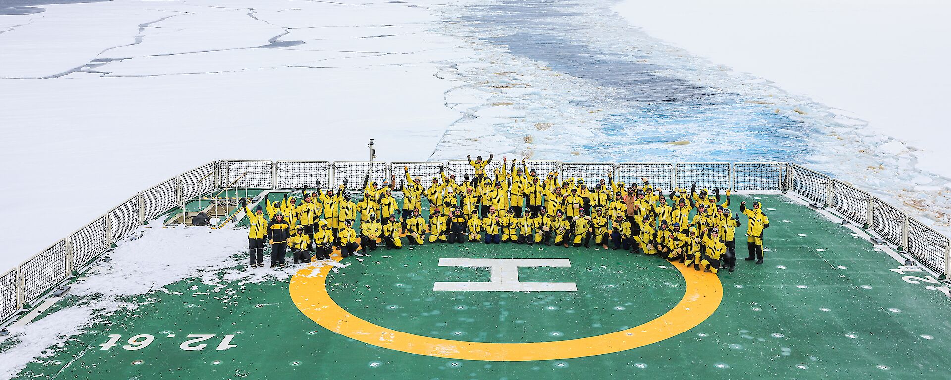 A large group of people in bright yellow cold weather jackets are posing for a group photo on the helicopter deck of an icebreaker ship.