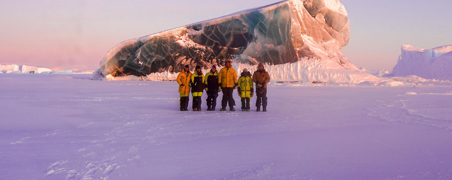 Six expeditioners at sunset with a jade iceberg in the distance.